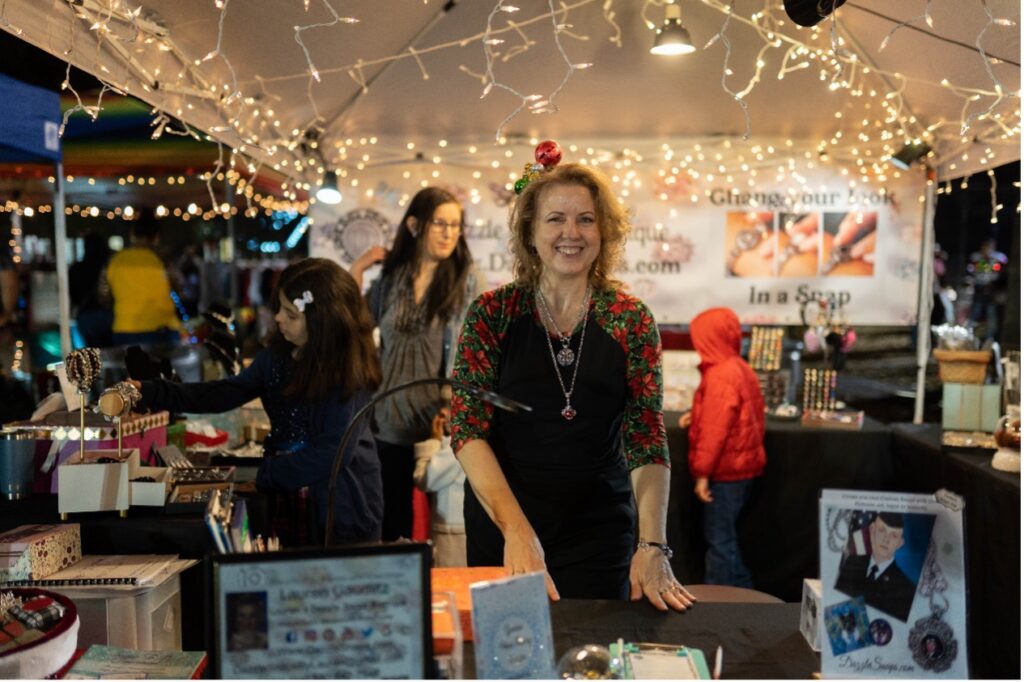 A woman stands behind a table dressed in festive Christmas gear near the vendor market. 