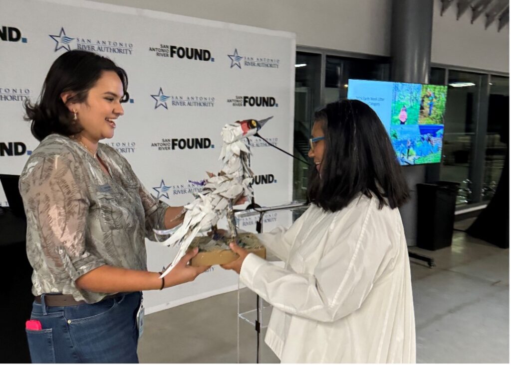 A woman hands another person a sculpture made out of trash in the shape of a whooping crane bird.