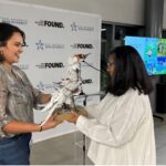 A woman hands another person a sculpture made out of trash in the shape of a whooping crane bird.